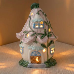 May include: A ceramic fairy house with a pink and blue floral design. The house has a flower-shaped roof, small windows, and a wooden door with a crescent moon. Green vines and leaves decorate the exterior. The windows and door glow with warm light.