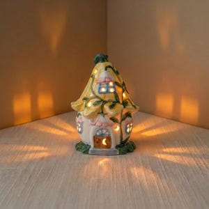 May include: A ceramic fairy house night light with a yellow roof and floral accents. The house has a small door and windows that emit a warm glow. The base is green, and the overall design is whimsical and decorative.