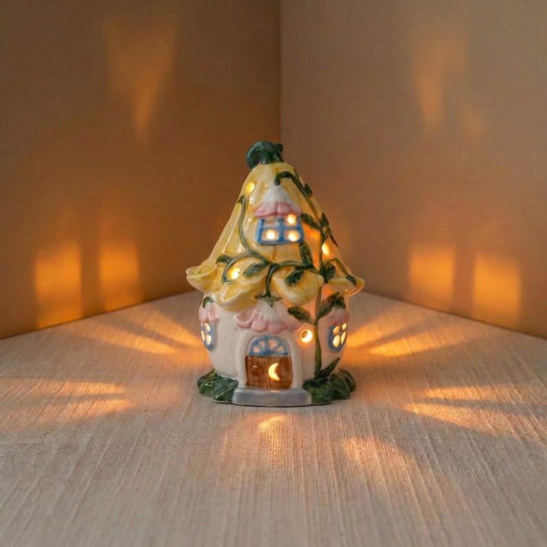 May include: A ceramic fairy house night light with a yellow roof and floral accents. The house has a small door and windows that emit a warm glow. The base is green, and the overall design is whimsical and decorative.