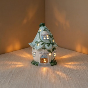 May include: A ceramic fairy house with a light blue roof and white walls. The house has several windows that glow with warm light. Green vines and leaves decorate the roof and walls. The door has a crescent moon design.