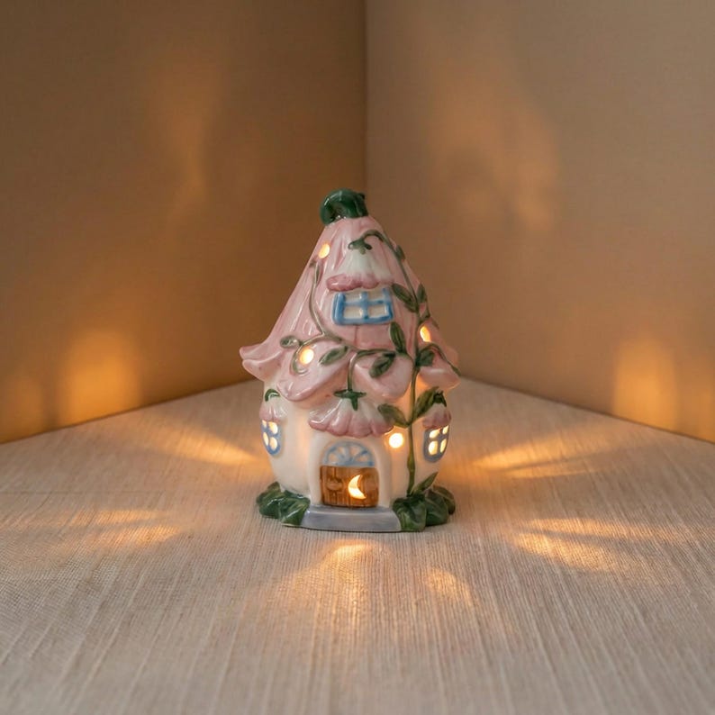 May include: A ceramic fairy house with a pink flower-shaped roof and a white base. The house has small windows, a wooden door with a crescent moon, and is adorned with green vines and leaves. The house is lit from within, creating a warm glow.
