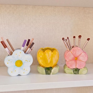 Op de afbeelding: Drie keramische bloemvormige containers. De eerste is wit met een gele kern, de tweede is geel en de derde is roze. Elk bevat make-up kwasten of potloden. De containers staan op een witte plank.