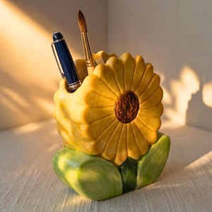 May include: A ceramic sunflower-shaped desk tidy with a pen and paintbrush inside. The organiser is yellow with a brown centre and green leaves at the base. The pen is blue and silver, and the paintbrush has a wooden handle.
