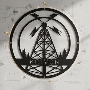 Peut inclure: Une décoration murale circulaire en métal noir représentant la silhouette d'une tour de transmission radio centrale avec des éclairs. Le design inclut de petits sapins à la base et le texte KC1VCN centré horizontalement.