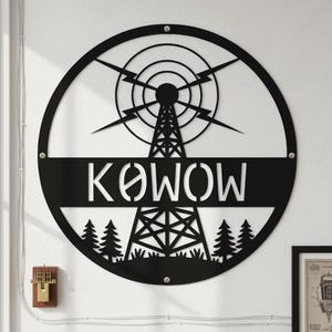 Puede incluir: Una pieza de arte mural circular de metal negro que presenta la silueta de una torre de transmisión de radio central, rayos estilizados y una hilera de pinos en la base. Una barra horizontal en el centro muestra el texto KOWOW en letras blancas.