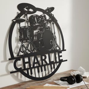 May include: Black metal drum kit wall art featuring the name "CHARLIE" and a piano keyboard design. The drum kit includes cymbals, drums, and stands. The art is displayed on a white wall, with headphones and drumsticks on a wooden surface.