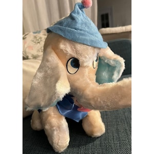May include: A plush toy elephant with large blue eyes, a long trunk, and large ears. The elephant is wearing a light blue nightcap with a pink pom-pom and a blue bow tie. The toy is cream-colored and has a soft, fuzzy texture.