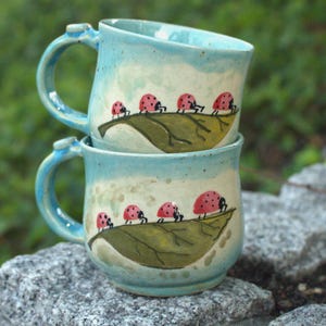 Peut inclure: Deux tasses en céramique empilées avec un vernis bleu clair et une finition mouchetée. Chaque tasse présente une feuille verte peinte à la main avec une rangée de coccinelles rouges à points noirs marchant sur la surface. Anse incurvée.