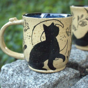 Puede incluir: Una taza de cerámica con esmalte crema moteado que presenta la silueta negra de un gato sentado frente a una luna creciente. El diseño incluye delicados dibujos de líneas negras de hojas y enredaderas. El interior de la taza es azul oscuro.
