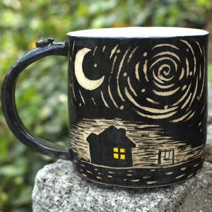 Può includere: Tazza in ceramica nera con un design a cielo notturno. La tazza presenta una luna crescente, stelle e una casa con una finestra gialla luminosa. Il manico è scuro e curvo. La tazza è appoggiata su una pietra grigia.