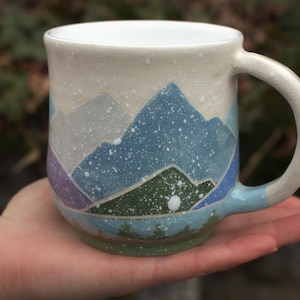 Può includere: Una tazza in ceramica con un design a paesaggio montano. La tazza presenta un interno e un manico bianchi, con un esterno beige chiaro. La scena della montagna include tonalità blu, verdi e viola, con macchie bianche. La tazza è tenuta in una mano.