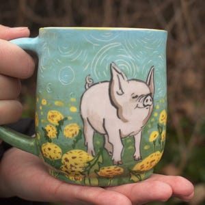 Könnte beinhalten: Keramikbecher mit hellblauem Hintergrund mit einer weißen Schweineillustration. Der Becher hat einen grünen Henkel und ein gelb-grünes Blumendesign. Der Becher wird in den Händen einer Person gehalten.