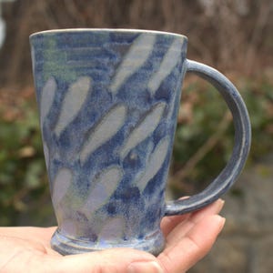 Könnte beinhalten: Blaue Keramiktasse mit einem weißen Muster aus länglichen Tropfen. Die Tasse hat einen großen Henkel und eine ausgestellte Form. Die Tasse wird in einer Hand gehalten. Die Tasse ist etwa 10 cm hoch.