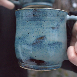 Op de afbeelding: Een handgemaakte keramische mok met een blauw glazuur. De mok heeft een afgeronde vorm met een oor en een gespikkelde textuur. De kleur gaat over van licht- naar donkerblauw, met bruine accenten. De mok wordt door een persoon vastgehouden.
