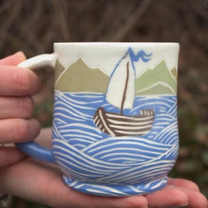 Könnte beinhalten: Eine Keramiktasse mit nautischem Thema. Die Tasse zeigt ein Segelboot auf blauen Wellen, mit grünen und beigen Bergen im Hintergrund. Der Griff und der Boden sind blau, und der Rand ist weiß.