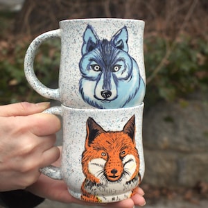Peut inclure: Deux mugs en céramique empilés, chacun avec une anse. Le mug supérieur présente un visage de loup bleu, et le mug inférieur a un visage de renard orange. Les deux mugs ont un fond blanc moucheté.