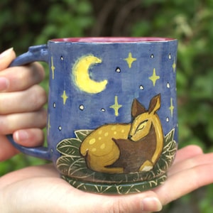 Könnte beinhalten: Blaue Keramik-Tasse mit handgemaltem Design eines schlafenden Hirsches unter einem gelben Halbmond und Sternen. Die Tasse hat einen dunkelblauen Henkel und eine rosa Innenseite. Die Tasse wird in den Händen einer Person gehalten.