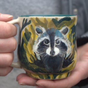May include: A ceramic mug with a hand-painted raccoon design. The raccoon has a black and white face with blue accents, surrounded by yellow and green foliage. The mug has a white handle and a teal interior.