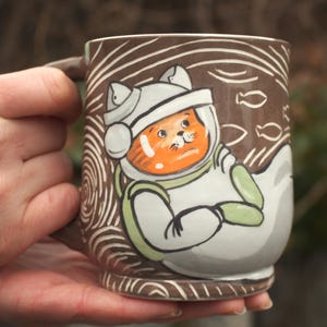 以下が含まれることがあります： 宇宙服を着たオレンジ色の猫のイラストが描かれた、茶色のセラミックマグカップ。マグカップには茶色の持ち手と、渦巻きと魚の装飾模様があります。