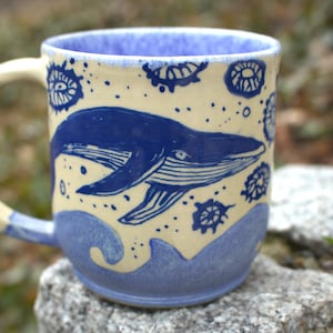 Può includere: Tazza in ceramica con un design a balena blu. La tazza presenta un corpo color crema con bordo, manico e motivo a onde blu alla base. La balena è raffigurata in blu, insieme ad altri elementi decorativi blu.