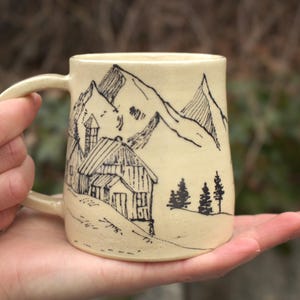 Könnte beinhalten: Cremefarbene Keramiktasse mit einer Schwarz-Tusche-Illustration einer Berglandschaft. Das Design zeigt eine Hütte, Berge und Bäume. Die Tasse hat einen gebogenen Henkel und wird in einer Hand gehalten.