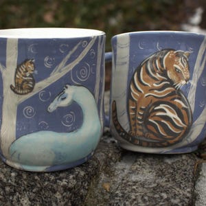 Może przedstawiać: Dwa ceramiczne kubki z niebieskimi uchwytami i niebieskim tłem. Jeden kubek przedstawia kota na drzewie i węża morskiego. Drugi kubek ma tygrysa. Kubki stoją na szarej skale.