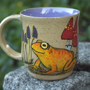 May include: A ceramic mug with a speckled beige exterior and a smooth lavender interior. The mug features a hand-painted illustration of a bright orange frog, several tall dark purple mushrooms, and two large red mushrooms with white spots.