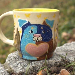 Puede incluir: Una taza de cerámica con un diseño caprichoso de perro. La taza tiene un exterior color crema y un interior amarillo. La ilustración del perro está dividida, con un lado azul y otro marrón, y un corazón rosa. Mariposas amarillas están dispersas alrededor de la taza.