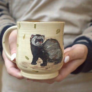 May include: A cream-coloured ceramic mug featuring a hand-painted illustration of a dark brown and black ferret standing on a patch of ground. The mug is decorated with small, scattered green leaf motifs and has a sturdy handle on the left side.