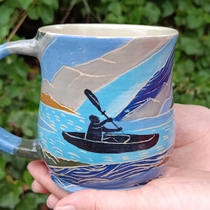 Peut inclure: Un mug en céramique orné d'une scène sculptée et peinte à la main représentant une personne dans un kayak noir pagayant sur une eau bleue. Le décor inclut des montagnes bleu clair en arrière-plan, des vagues texturées et un bord intérieur clair.