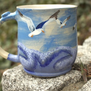 Op de afbeelding: Keramische mok met een handgeschilderd ontwerp van zeemeeuwen in vlucht boven een blauw-witte oceaanscène. De mok heeft een lichtblauw oor en een crèmekleurig interieur. De mok staat op een grijze steen.