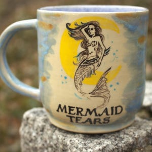 May include: A white ceramic mug with a black handle and the text "But First, Coffee" in a bold, black font. The mug is on a wooden surface. The mug is approximately 10 cm tall.