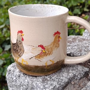 Pode incluir: Uma caneca de cerâmica com esmalte bege claro apresentando uma cena pintada à mão de três galinhas em tons de castanho, branco e dourado. As galinhas têm cristas vermelhas e bicos amarelos, posicionadas sobre uma base castanha escura.