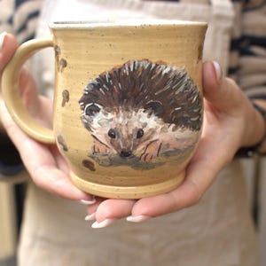May include: A ceramic mug with a speckled tan glaze featuring a hand-painted illustration of a brown and white hedgehog on the front. The mug has a rounded body, a sturdy handle on the left side, and a slightly flared rim with a smooth finish.
