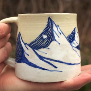 May include: Cream-colored ceramic mug with a blue mountain range design. The mountains are depicted with white and blue brushstrokes. The mug has a curved handle and is held in a person's hand.
