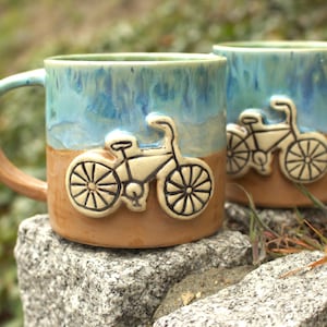 Op de afbeelding: Twee keramische mokken met een fietsreliëf ontwerp. De mokken hebben een turquoise en bruin kleurenschema. Het fietsontwerp is beige. De handgreep van één mok is bruin. De mokken staan op een grijze steen.