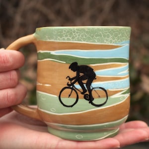 Könnte beinhalten: Eine Keramiktasse mit einer Radfahrer-Silhouette. Die Tasse hat einen beigefarbenen Henkel und ein gemaltes Landschaftsdesign in Grün, Blau und Beige. Die Tasse wird von einer Person gehalten.