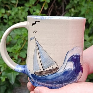 Könnte beinhalten: Ein cremefarbener Keramikbecher mit einer handgemalten Szene eines Segelboots auf einer dunkelblauen Meereswelle. Das Design zeigt zwei schwarze Vögel, die am Himmel über dem Boot fliegen. Der Becher verfügt über einen geschwungenen Henkel.