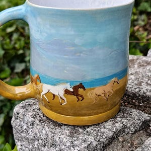 Peut inclure: Une tasse en céramique ornée d'une scène peinte à la main représentant des chevaux galopant sur un paysage ocre sous un ciel bleu clair. La tasse présente une finition lisse et brillante, une anse incurvée et un intérieur blanc.
