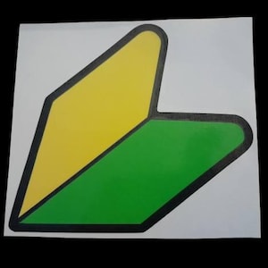 May include: A sticker featuring the Japanese beginner driver symbol, a yellow and green stylized arrow shape with a black outline. The sticker is on a white background.