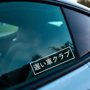 May include: White rectangular sticker with Japanese text, "遅い車クラブ", on a car window. The sticker is in a white rectangular frame. The car window is tinted and reflects the surroundings.
