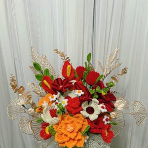 Puede incluir: Un vibrante arreglo floral con flores de ganchillo hechas a mano en tonos rojos, naranjas, blancos y verdes. El ramo incluye varios tipos de flores, con acentos decorativos y follaje dorados. El arreglo se presenta en un jarrón gris.