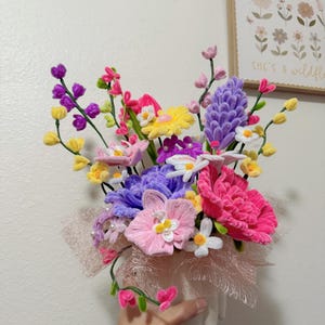 May include: A vibrant handmade flower arrangement in a white vase. The bouquet features a variety of colourful flowers in shades of pink, purple, yellow, and white. The arrangement is accented with a pink ribbon and held by a hand.