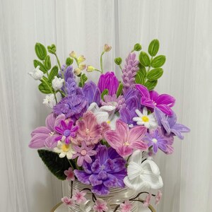 May include: A colourful floral arrangement in a white woven basket. The bouquet features plush flowers in shades of purple, pink, and white, with green foliage. The arrangement is set on a gold-toned table.