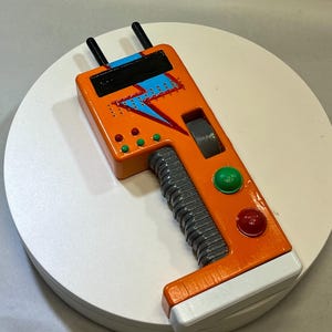 May include: A handheld orange device featuring a blue lightning bolt graphic, two black antennas, a grey ribbed handle, and multiple circular buttons in red, green, and grey. The rectangular unit includes a black screen area and a white base section.