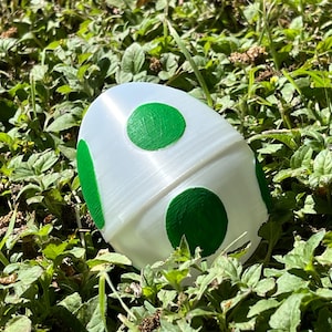 May include: A white, egg-shaped object with green circular and oblong markings, resting in a bed of green foliage. The object has a textured surface and appears to be a decorative item or toy. The design is reminiscent of a video game character.