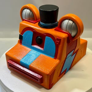 May include: A 3D printed orange camera-shaped object featuring a black top hat, two rounded ears with silver accents, and bright blue rectangular details. The front includes a central black lens, a red horizontal slot, and a blue lightning bolt.