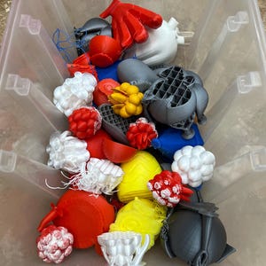 Puede incluir: Un contenedor de plástico transparente lleno de varios objetos impresos en 3D en rojo, blanco, amarillo, gris y azul. Los objetos incluyen un par de guantes rojos, esferas y otras formas abstractas. El contenedor está sobre una superficie marrón.