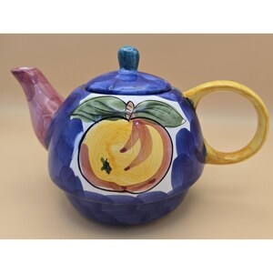 Puede incluir: Una tetera de cerámica con un diseño pintado a mano de un melocotón amarillo con hojas verdes sobre un fondo azul intenso. La tetera presenta un pico en tonos rosados, un asa amarilla y una tapa azul a juego con un pequeño pomo.