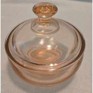 May include: A small, round, light pink glass dish with a matching removable lid. The dish features a smooth, translucent finish and a central knob handle on the lid. The glass has a subtle peach-toned hue and a simple, circular design.